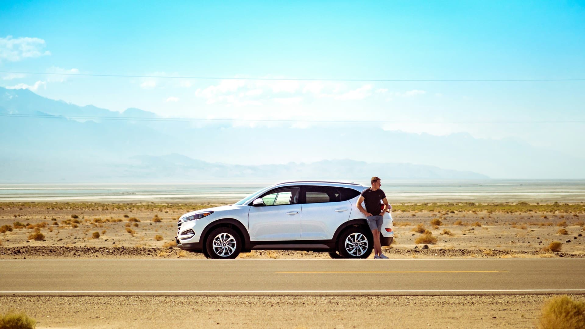 A man leans against a white SUV parked on the gravel edge of an empty desert road, with a pale valley floor and hazy mountains behind him.