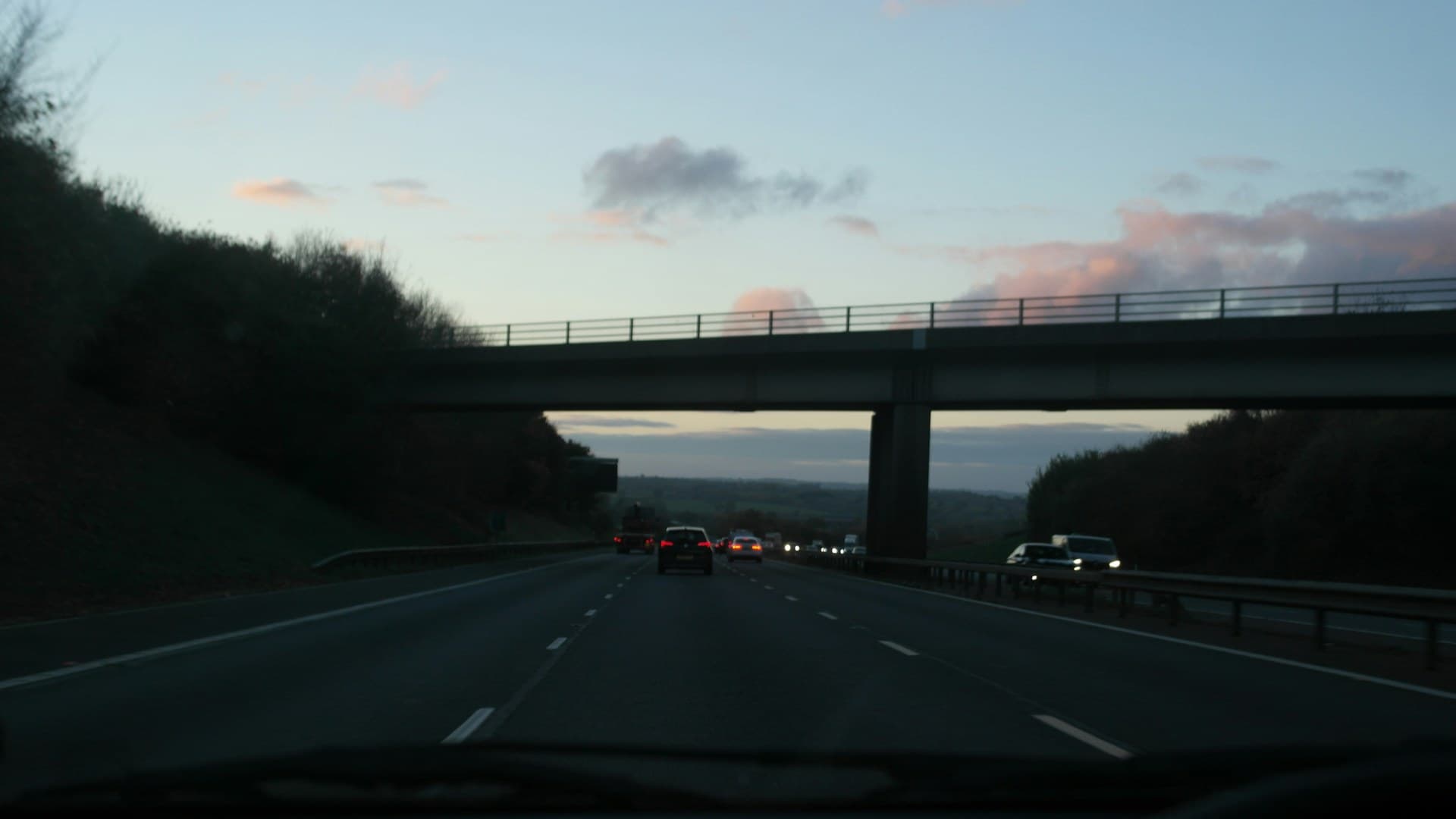 The view through a windscreen on a three-lane motorway at dusk: tail lights ahead, a concrete bridge overhead and a pale evening sky with pink clouds.